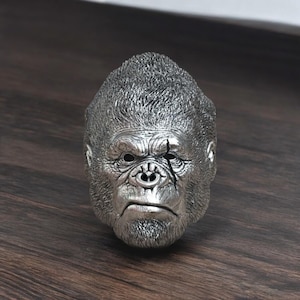 Puede incluir: Un anillo de cabeza de gorila de plata con un acabado texturizado detallado. La cara del gorila tiene una expresión seria y una cicatriz sobre el ojo.