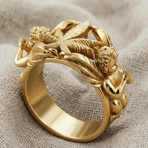 May include: A gold-colored ring featuring detailed cherub figures and a central cannabis leaf design. The ring has an ornate, sculpted appearance, with the cherubs in a praying pose. The ring is made of a shiny, metallic material.
