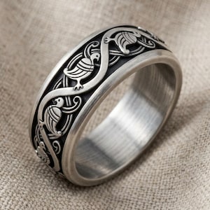 May include: A silver-toned metal ring featuring a continuous embossed pattern of stylized birds intertwined with flowing, curved lines. The background of the band is darkened to highlight the raised, polished silver bird motifs and scrollwork.