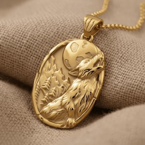 Può includere: Un ciondolo ovale color oro su catenina, caratterizzato da un design in rilievo che raffigura un lupo che ulula alla luna piena. La scena include cime montuose stilizzate e alberi sempreverdi sullo sfondo. Il ciondolo è in metallo dorato.
