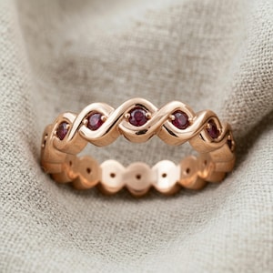 Peut inclure: Bague en or rose avec un motif ondulé continu, sertie de petites pierres précieuses rouges rondes. La bague a une finition polie et est présentée sur un tissu de couleur neutre. Les pierres précieuses sont espacées uniformément.
