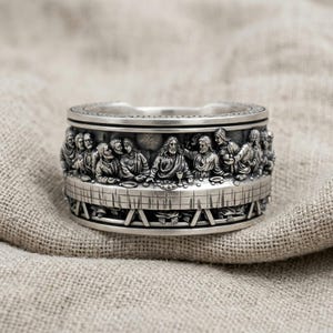 Può includere: Un anello a fascia larga in argento con un dettagliato rilievo tridimensionale dell'Ultima Cena. Il design raffigura diverse figure sedute a un lungo tavolo, realizzate in argento lucido con aree incavate scure per evidenziare gli intagli.