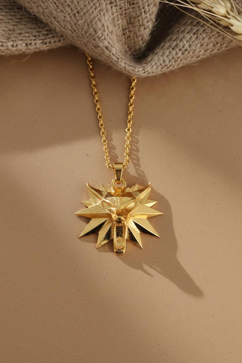 May include: A gold-colored pendant necklace featuring a stylized wolf head emblem. The pendant is intricately designed with sharp, angular details and is suspended from a delicate gold chain. The necklace is displayed against a neutral background.