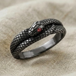 Puede incluir: Un anillo de metal gris oscuro con forma de serpiente enroscada y un patrón de escamas texturizado que cubre la banda. La cabeza de la serpiente presenta una pequeña piedra preciosa roja redonda y facetada como ojo, con un aspecto detallado.