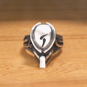 Puede incluir: Un anillo de plata con un diseño inspirado en Predator. El anillo presenta una cabeza detallada del Predator con un símbolo en relieve en la frente.