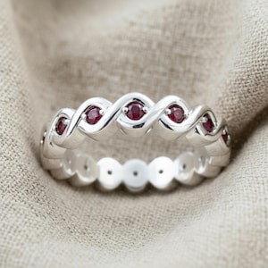 Puede incluir: Un anillo de tono plateado con un diseño de banda de metal trenzada continua. El anillo está engastado con múltiples piedras preciosas pequeñas, redondas y de color rojo intenso incrustadas en las curvas de la estructura metálica.