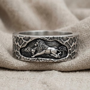 May include: A silver-toned metal ring featuring a detailed relief carving of a lion in a savanna landscape. The band is decorated with a textured, irregular pattern resembling giraffe spots, creating a contrast between the smooth lion and the background.