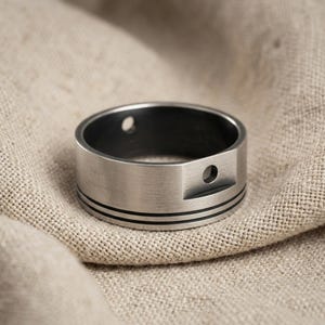 May include: A brushed silver-tone metal ring featuring a wide band with two thin horizontal black engraved lines circling the exterior. The ring includes a raised section with a small circular hole and an additional small hole on the band.