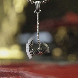 May include: A silver-colored pendant necklace featuring a detailed axe pendant. The axe head has intricate designs and a double-bladed edge. The pendant hangs from a delicate chain, creating a striking accessory.