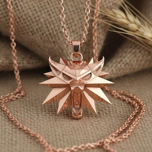 May include: Rose gold pendant necklace featuring a stylized wolf head emblem. The pendant is intricately designed with sharp angles and a detailed face, suspended from a delicate chain. The necklace is displayed on a neutral, textured background.