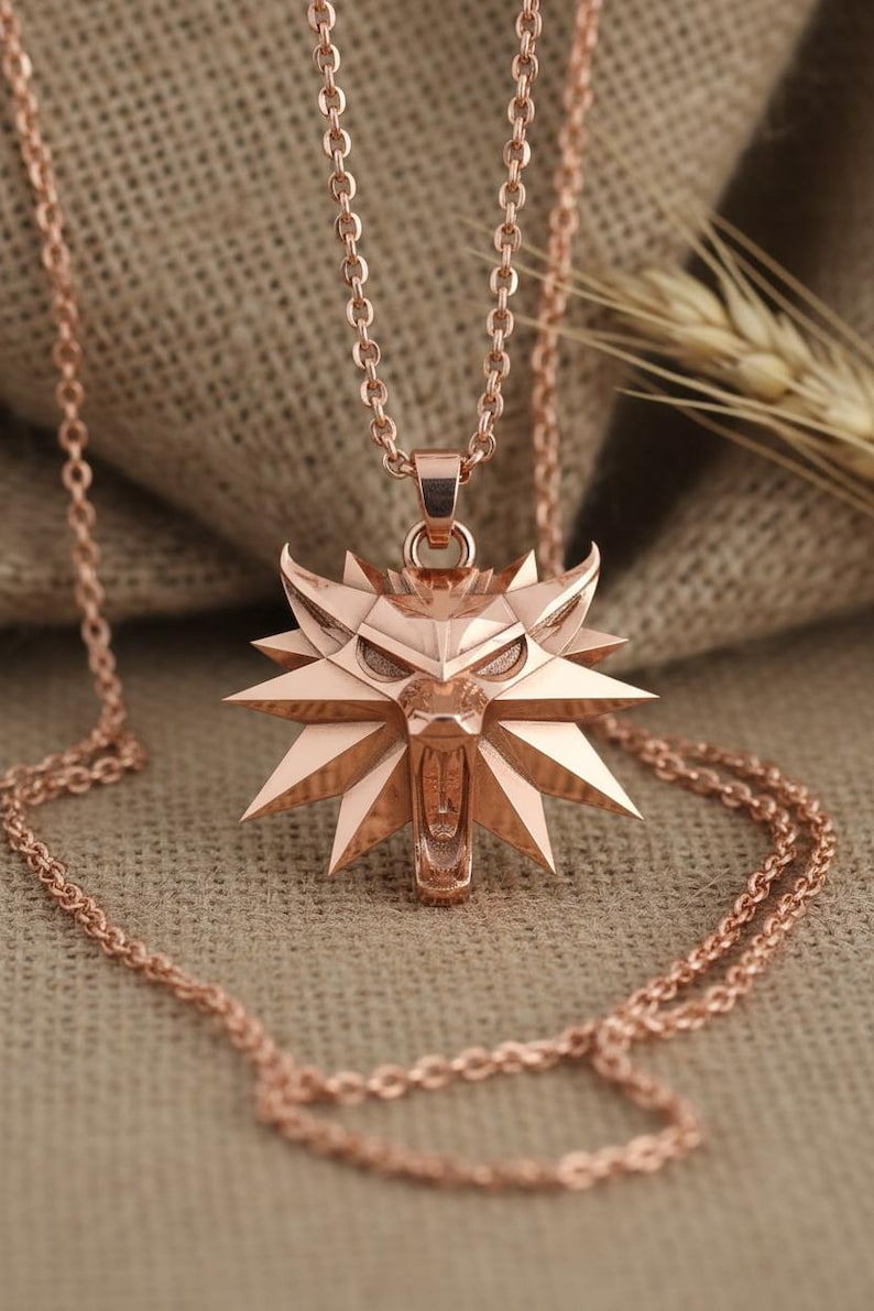 May include: Rose gold pendant necklace featuring a stylized wolf head emblem. The pendant is intricately designed with sharp angles and a detailed face, suspended from a delicate chain. The necklace is displayed on a neutral, textured background.