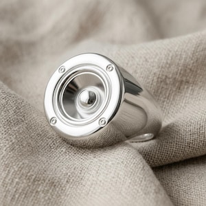 A silver ring with a circular design resembling a speaker or microphone is placed on a beige fabric background.