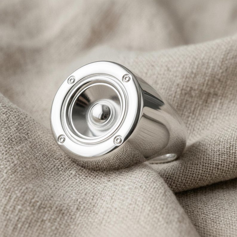 A silver ring with a circular design resembling a speaker or microphone is placed on a beige fabric background.