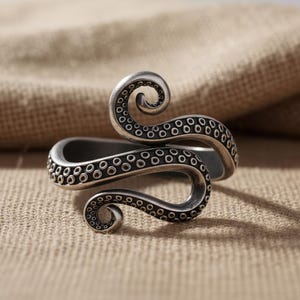 May include: A silver-toned ring featuring an octopus tentacle design. The ring wraps around the finger with two curling tentacles, each detailed with small suction cup markings. The ring has a vintage or antique appearance.