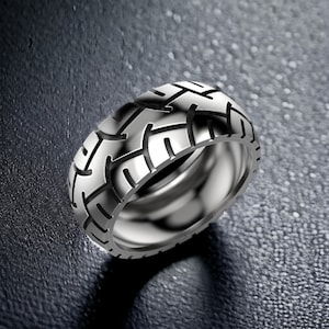 Może przedstawiać: Srebrny pierścionek z wzorem bieżnika opony. Pierścionek jest wykonany z metalu i ma błyszczące wykończenie.