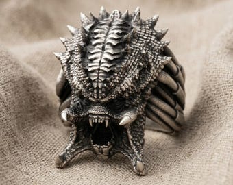 May include: A silver-toned ring featuring a detailed, textured creature head with a wide-open mouth, sharp fangs, and multiple spikes along the top. The design includes intricate scales and ridges, with a dark, oxidized finish highlighting the details.