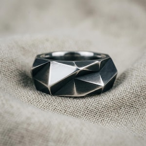 Könnte beinhalten: Ein dunkelgrauer Metallring mit einer facettierten, geometrischen Oberfläche und scharfen, kantigen Ebenen. Das Band hat ein gebürstetes, mattes Finish, das die komplexe, vielseitige Struktur des Designs hervorhebt und ein Spiel von Licht und Schatten erzeugt.