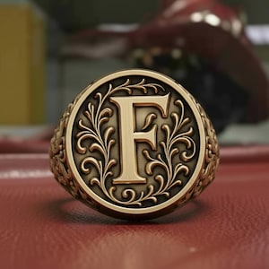 Puede incluir: Un anillo dorado con la letra "F" en el centro, rodeado de un diseño floral ornamentado. El anillo tiene una banda detallada y texturizada. El fondo es una superficie roja borrosa.