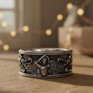 May include: A silver ring with an intricate design featuring an angel with a sword. The ring has a wide band with detailed scrollwork and a central angel figure. The ring is displayed on a light brown surface.