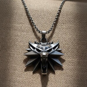 May include: A silver-colored pendant necklace featuring a stylized wolf head emblem. The pendant has a detailed, angular design with sharp edges and a fierce expression. The chain is a linked design, and the background is a textured, neutral fabric.