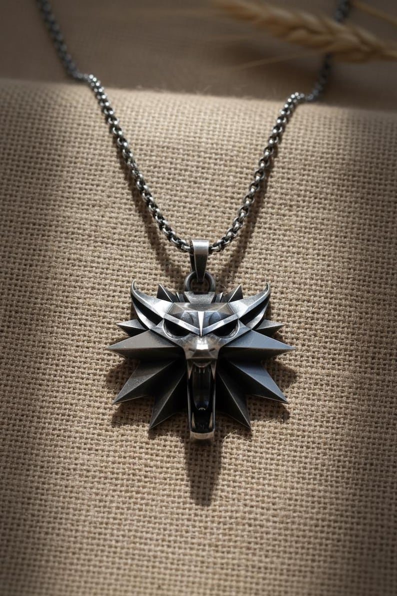 May include: A silver-colored pendant necklace featuring a stylized wolf head emblem. The pendant has a detailed, angular design with sharp edges and a fierce expression. The chain is a linked design, and the background is a textured, neutral fabric.