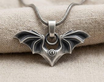 May include: A silver-toned metal pendant shaped like bat wings with a central eye-like detail, suspended from a matching snake chain. The pendant features a dark, oxidized finish in the recessed areas of the wings and a smooth, polished bail.