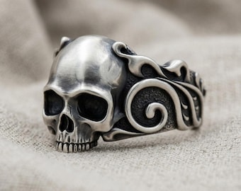 Puede incluir: Un anillo de plata que presenta un diseño de calavera detallado como pieza central. La banda está elaborada con intrincados patrones de volutas y espirales en relieve que se extienden desde los lados de la calavera, con un acabado pulido.