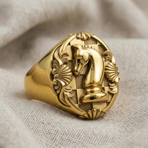 May include: A gold-colored ring featuring a chess knight design. The ring has an oval face with a checkered background and ornate detailing. The knight is in the center, surrounded by decorative elements.