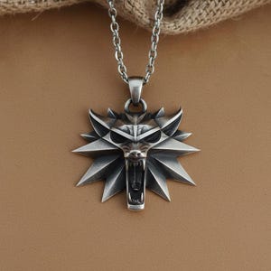 May include: A silver-colored pendant necklace featuring a wolf's head emblem with an open mouth and sharp teeth. The pendant is attached to a chain necklace. The background is a neutral brown.