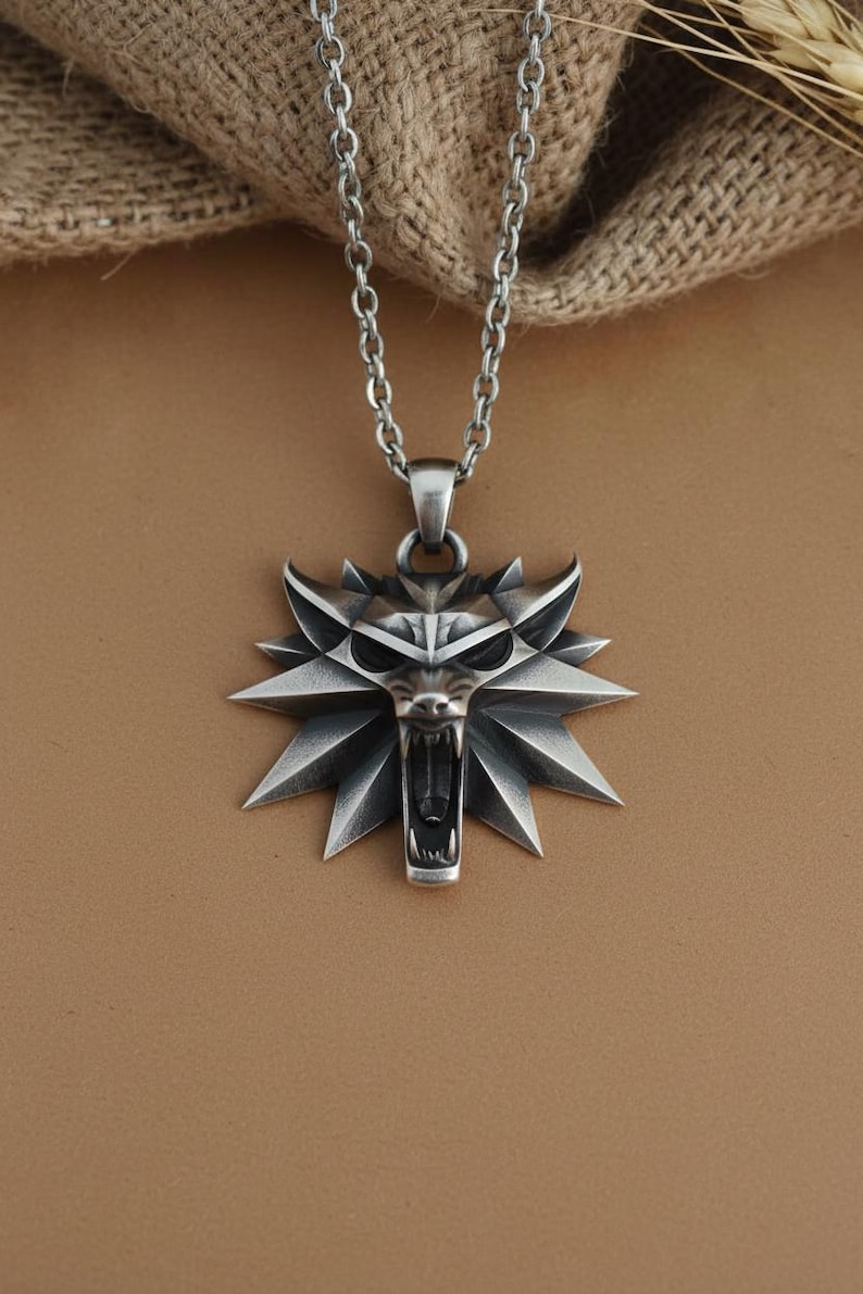 May include: A silver-colored pendant necklace featuring a wolf's head emblem with an open mouth and sharp teeth. The pendant is attached to a chain necklace. The background is a neutral brown.