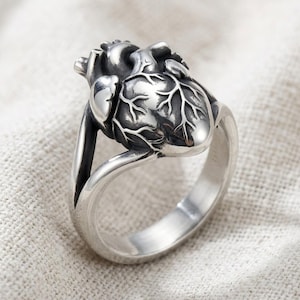 Puede incluir: Un anillo de tono plateado que presenta una escultura tridimensional detallada de un corazón anatómico como pieza central. La superficie metálica tiene un acabado pulido con áreas hundidas oscurecidas para resaltar las venas y la estructura.