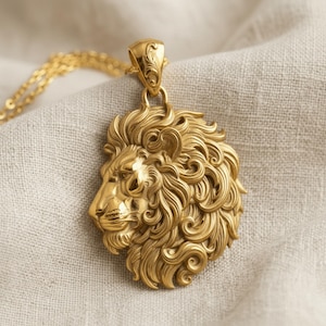 Puede incluir: Un colgante de tono dorado que presenta una cabeza de león detallada de perfil con una melena fluida y rizada. El colgante está unido a una cadena de eslabones dorada a juego, mostrando un trabajo de metal intrincado y un acabado pulido.