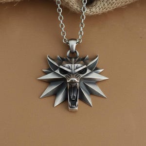 May include: A silver-colored pendant necklace featuring a stylized wolf head emblem. The pendant has a detailed, angular design with an open mouth and sharp teeth. The necklace chain is also silver and has a link design. The background is a neutral brown.