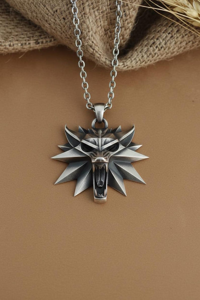 May include: A silver-colored pendant necklace featuring a stylized wolf head emblem. The pendant has a detailed, angular design with an open mouth and sharp teeth. The necklace chain is also silver and has a link design. The background is a neutral brown.