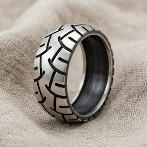 May include: A silver-toned metal ring featuring a textured exterior pattern that mimics the tread of a tyre. The band has a brushed finish on the outer surface with deep, dark grooves forming the pattern, while the interior is smooth and dark.
