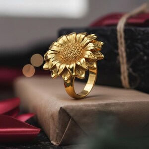 Puede incluir: Un anillo dorado con un diseño detallado de girasol. Los pétalos y el centro del anillo están intrincadamente elaborados, mostrando una apariencia floral realista. El anillo se exhibe sobre una caja de regalo marrón, con un fondo borroso.