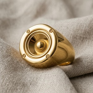 A gold-colored ring with a speaker design sits on a beige fabric surface.