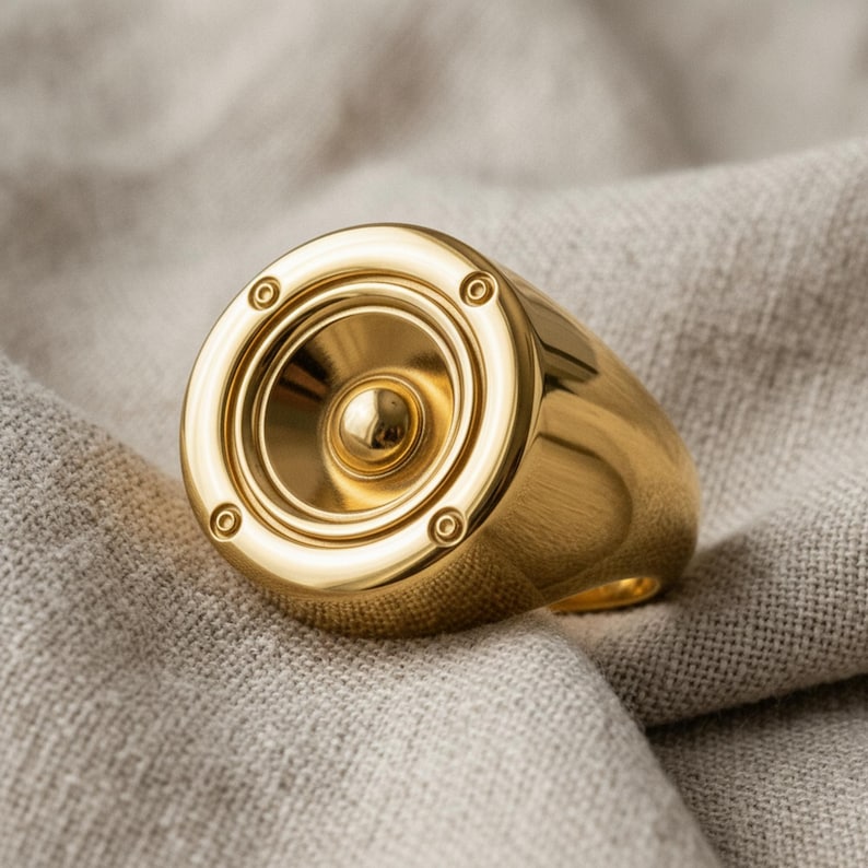 A gold-colored ring with a speaker design sits on a beige fabric surface.