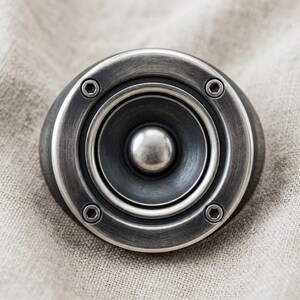 A close-up image of a silver-colored speaker with a circular opening in the center, resting on a textured fabric background.