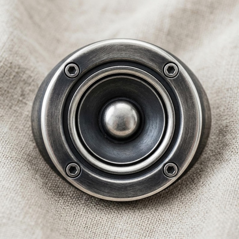 A close-up image of a silver-colored speaker with a circular opening in the center, resting on a textured fabric background.