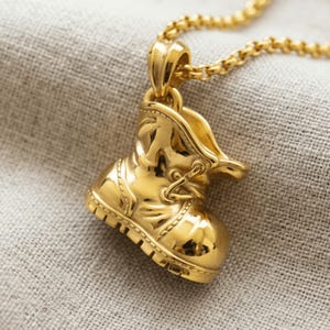 May include: A gold-toned metal pendant shaped like a small work boot, suspended from a matching gold-toned chain. The pendant features detailed molded textures representing laces, eyelets, and a thick lug sole, with a polished, reflective finish.