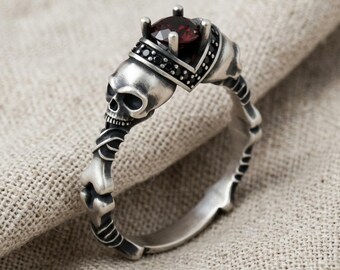 Puede incluir: Un anillo de plata con una piedra preciosa de color rojo oscuro en el centro, sujeta por cuatro garras. La banda presenta un diseño de calavera en la parte superior y una estructura ósea en los laterales, con pequeñas piedras negras.