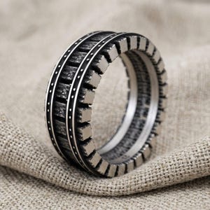 Puede incluir: Un anillo de tono plateado que presenta una banda central texturizada y oscurecida con un patrón de bloques repetitivos. El anillo está enmarcado por dos rieles plateados delgados y lisos con detalles de puntos espaciados en los bordes.