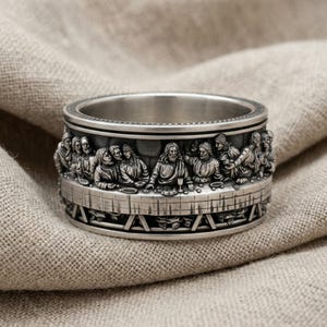 Può includere: Un anello in metallo color argento caratterizzato da un dettagliato rilievo scultoreo della scena dell'Ultima Cena che avvolge l'intera fascia. Il design raffigura figure sedute a un lungo tavolo, con finitura metallica e zone incavate.