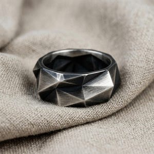 Op de afbeelding: Een donkergrijze metalen ring met een gefacetteerd, geometrisch oppervlak en een geborstelde afwerking. De band heeft een meerzijdig, hoekig ontwerp dat een spel van licht en schaduw creëert op de metalen textuur, wat zorgt voor een scherpe uitstraling.
