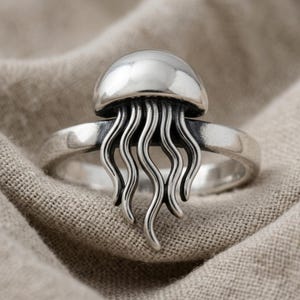 May include: A polished silver ring featuring a three-dimensional jellyfish design. The ring has a smooth, rounded dome representing the jellyfish bell, with several slender, wavy tentacles extending downward over the front of the band.
