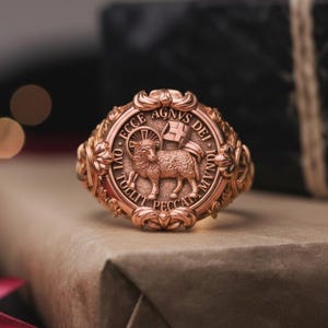 May include: A detailed rose gold ring with a religious motif, featuring a lamb and text. The ring is set on a wrapped gift box, with a blurred background. The ring's design is intricate and ornate.