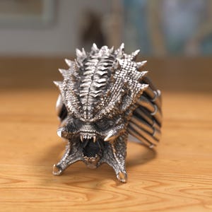 May include: A silver Predator head ring with a detailed design. The ring features a textured, scaly head with sharp teeth and a menacing expression.