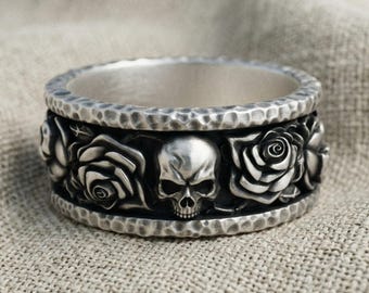 Könnte beinhalten: Ein silberner Ring mit einem Mittelband, das ein Reliefmuster aus Totenköpfen und Rosen zeigt. Die oberen und unteren Kanten des Rings haben eine gehämmerte, texturierte Oberfläche. Das Metall wirkt in den Vertiefungen dunkel und oxidiert.