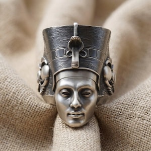 May include: A silver-toned ring featuring a detailed sculptural face of an Egyptian figure wearing a tall headdress with a central cobra emblem. The metal has a dark, oxidized finish in the recessed areas, highlighting the facial features.